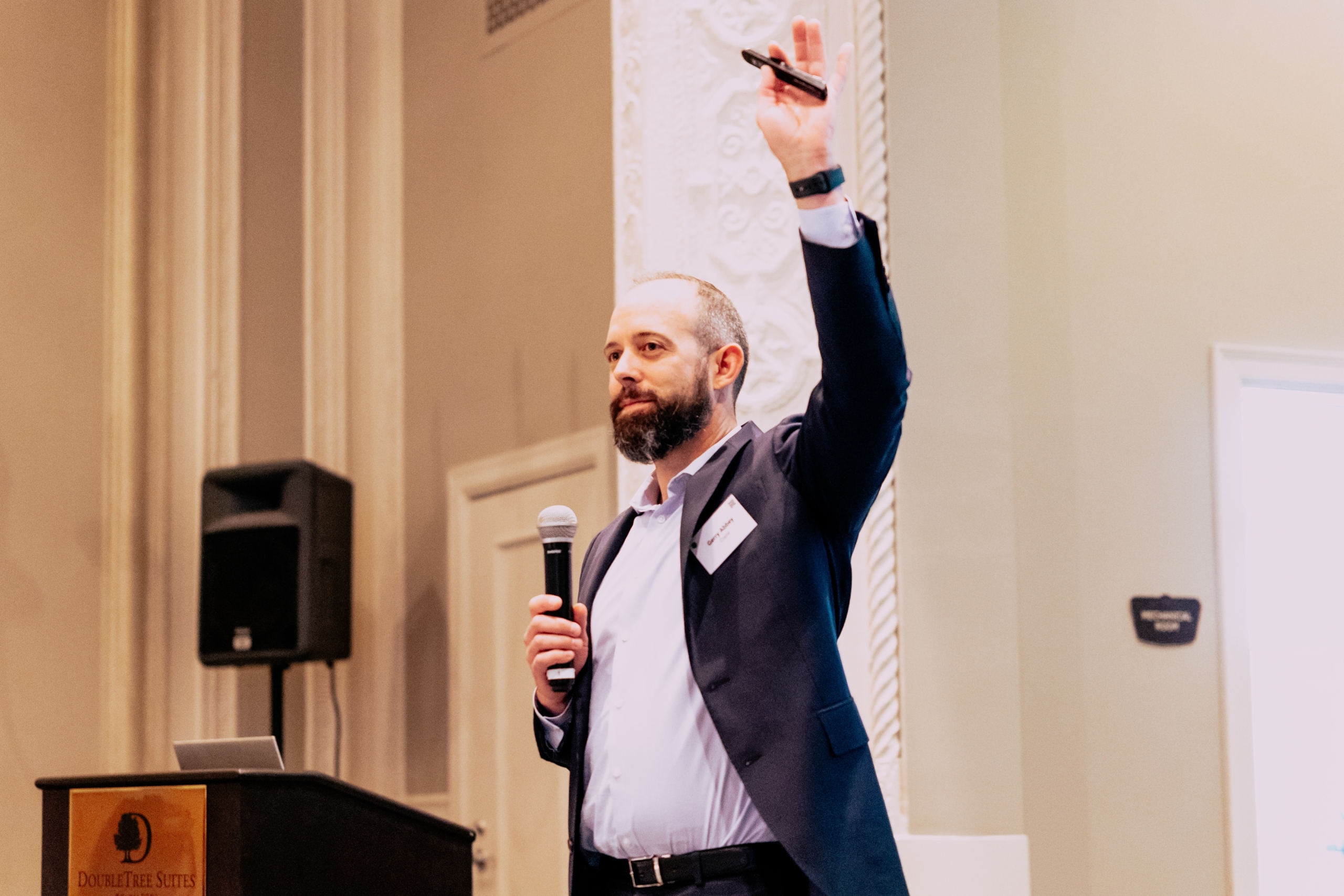 A man in a suit holding a microphone and raising his hand during a presentation at a conference.