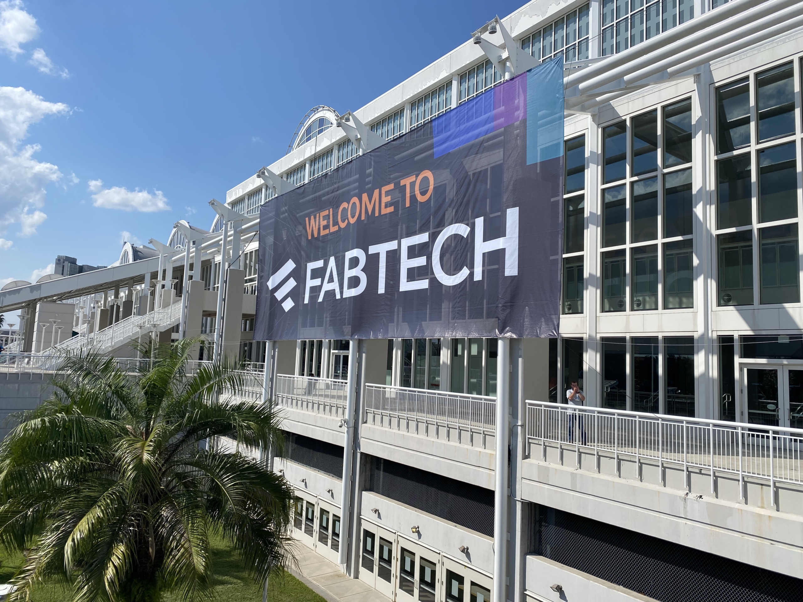 A large banner reading "WELCOME TO FABTECH" hangs outside a modern glass building under a blue sky.