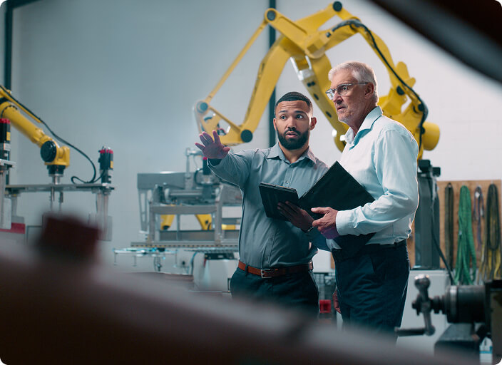 Two controls engineers discuss machinery in a factory with robotic arms and industrial equipment in the background.