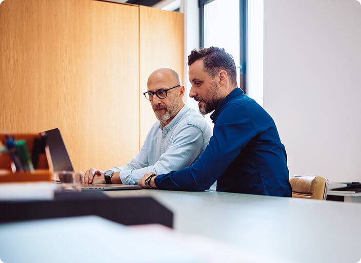 Two IT professionals and CISOs collaborate at a desk in a modern office, reviewing data on a laptop.